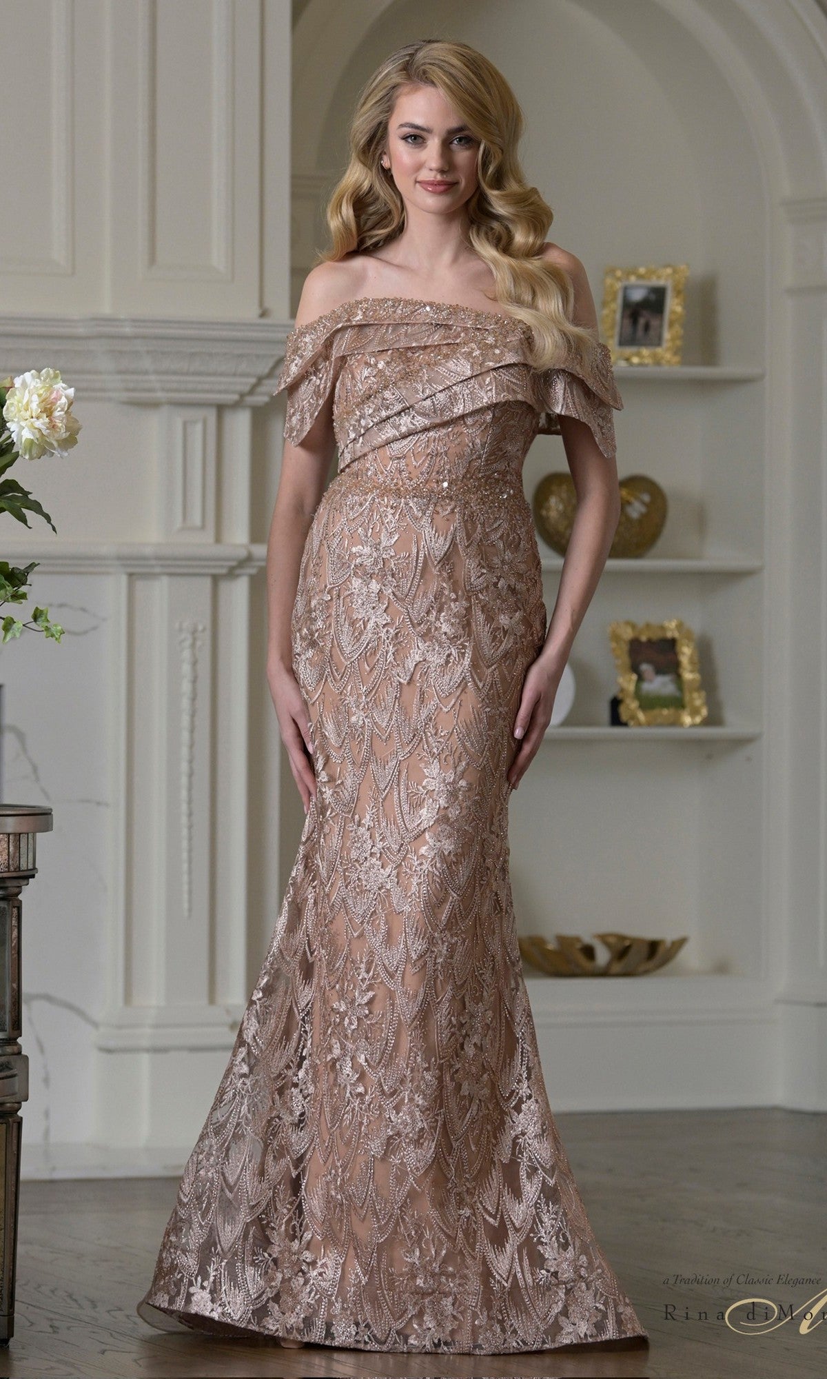 A woman stands indoors in the Rina Di Montella RD3531, a floor-length, off-the-shoulder rose gold dress with lace and metallic embroidery—an elegant mother-of-the-bride choice; decor shelves appear in the background.
